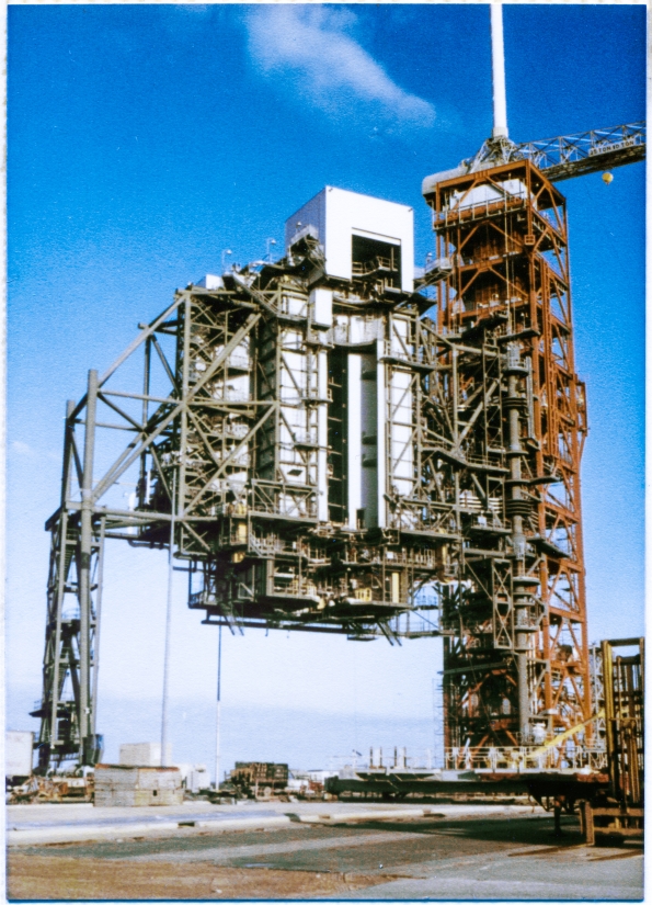 Image 047. We are drawing toward the close of contract NAS10-9655, and most of the steel on Drawing 79K14110  MODIFICATIONS TO LAUNCH PAD 39B FOR SPACE SHUTTLE – TASK II, under Specification 79K14111 at the John F Kennedy Space Center, Florida, has been furnished and installed, but we’re not quite there just yet. The Rotating Service Structure has yet to move. Has yet to demonstrate that it can roll its 4 million pound weight along the pair of curved steel rails which support it, allowing it to cross the open gulf of the Flame Trench on the Rail Beam which bridges the open gap, and continue on, until it spans the full width of the Flame Trench, sitting at right angles to its length, locked down in the Mate position it will occupy when there is a Space Shuttle on the Pad, requiring pre-launch servicing, before it flies. You are looking at both the RSS and the FSS from a viewpoint south of the Flame Trench, a bit east of the easternmost of the two widely-spaced double-Trackways which the treads of the Crawler will bear down on, as it carries the MLP to its launch position before the RSS is rotated around, and mated to the Shuttle which stands on top of it. Soon, the RSS will move. But not just yet. Not today. Photo by James MacLaren.